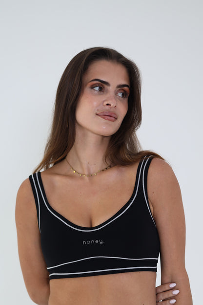 A woman wearing a black sports bra with white trim and the brand name 'honey.' printed on the front, photographed against a plain light gray background.